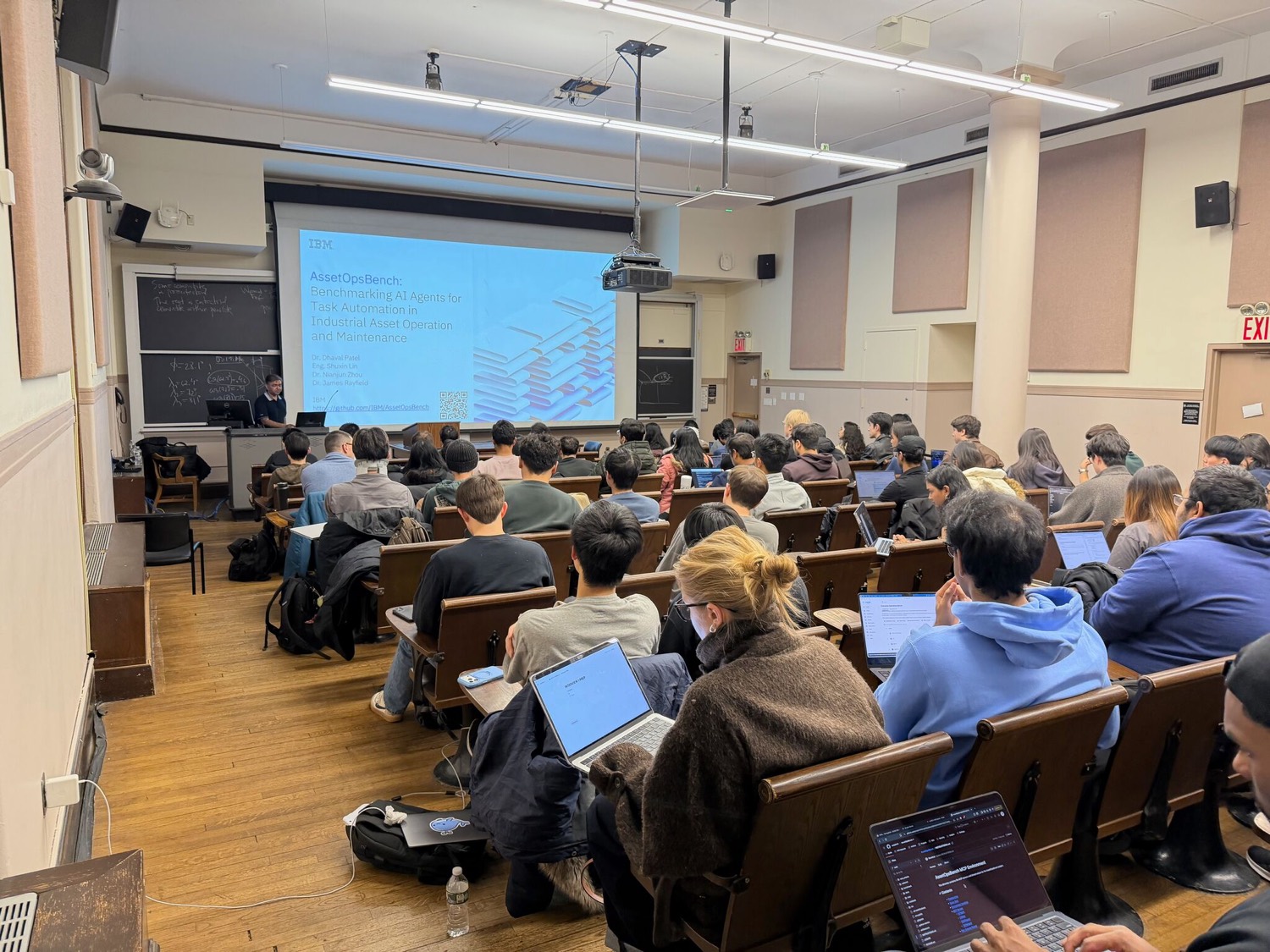 Dr. Dhaval Patel presenting the AssetOpsBench title slide to a full Columbia HPML lecture hall, with students following along on laptops.
