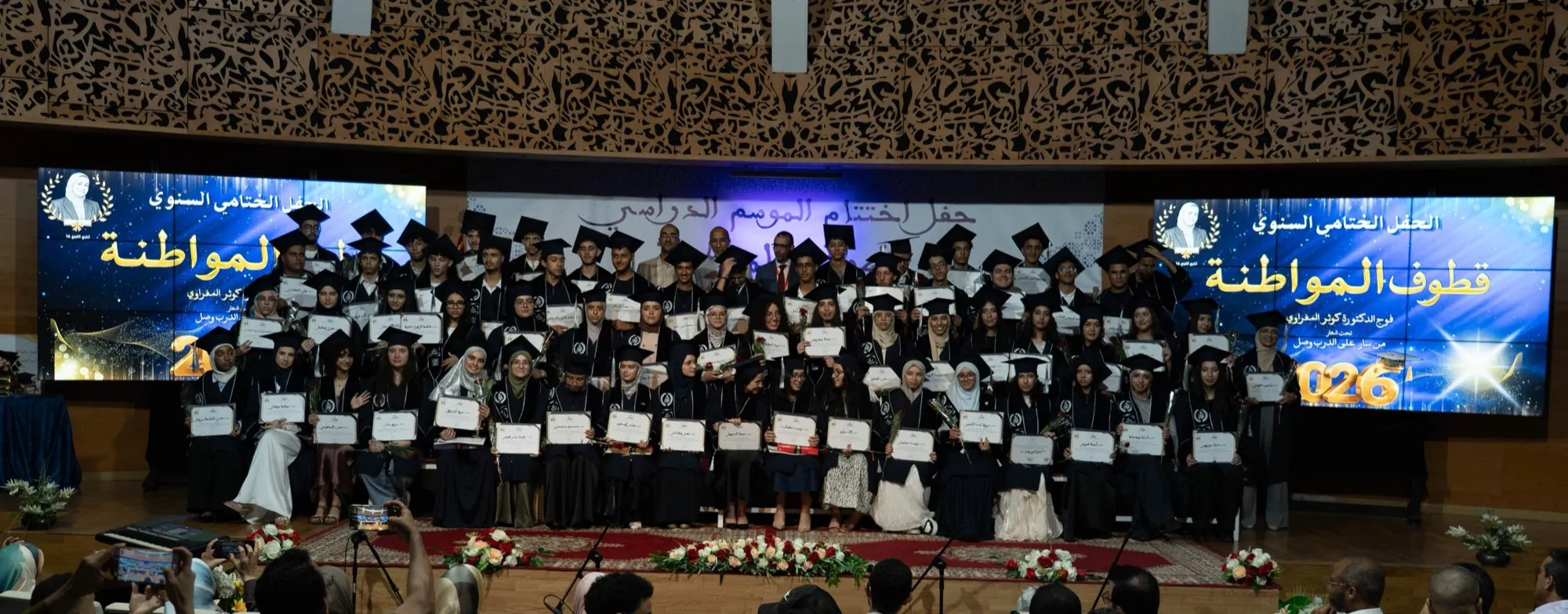 The 16th Baccalaureate graduating cohort of Groupe Scolaire Al Mouwatana in caps and gowns on stage, holding their diplomas